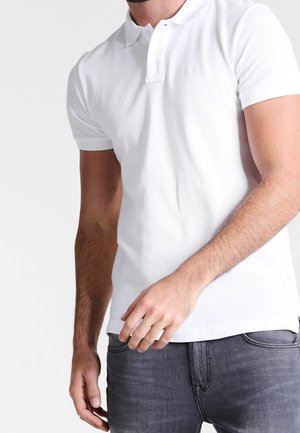 Homme portant un polo blanc cintré à manches courtes et un jean gris foncé, debout devant un fond clair uni.