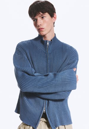 Jeune homme aux cheveux courts et foncés portant un pull zippé côtelé bleu, les bras croisés et regardant sur le côté devant un fond uni.