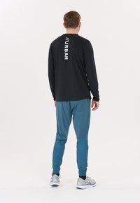 Svart långärmad tröja med vit "//URBAN" text på baksidan, tillsammans med turkosa träningsbyxor och grå löparskor. Enfärgad bakgrund.