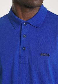 Modrá polo košile vyrobená z hladké tkaniny, s klasickým límečkem, třemi knoflíky a vyšitým logem "BOSS" v černé barvě na hrudi.