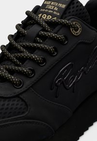 Μαύρο αθλητικό παπούτσι από υλικό με υφή και διατρήσεις, με κορδόνια με κίτρινα σημεία και ανάγλυφη λεπτομέρεια λογότυπου στο πλάι.