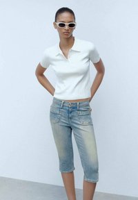 Polo shirt bianco a costine, jeans capri in denim blu chiaro con sezioni sbiadite e tasche frontali, abbinati a occhiali da sole ovali bianchi.