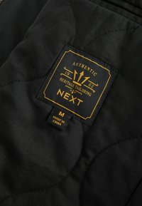 Tissu matelassé noir avec une étiquette cousue indiquant "Authentic Heritage Tailoring NEXT 1982" et une étiquette de taille marquée Medium, fabriqué en Chine.