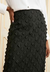 Jupe noire avec un motif floral texturé et des accents en relief. Tissu lisse avec un design ajusté, portée avec un haut blanc à manches longues.