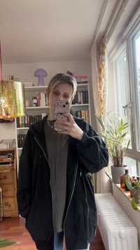 Zwarte oversized jas met een ritssluiting aan de voorkant, losvallend model en grote zakken, gedragen over een grijs shirt, met een simpele zilveren ketting.