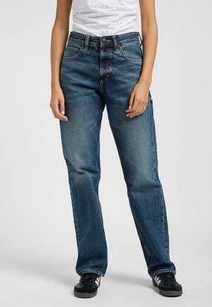 Jean en denim bleu taille haute avec une coupe droite, texture délavée, cinq poches et détails de couture dorés. Associé à des baskets noires.