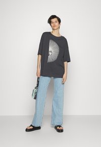 Tmavě šedé oversized tričko s bílou grafikou slunce, spárované se světle modrými širokými džíny a černými sandály. Součástí je malá vzorovaná taška.