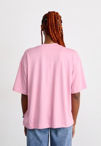 Personne avec de longues nattes rouges portant un T-shirt ample rose clair et un jean bleu, se tenant de dos devant un fond uni.