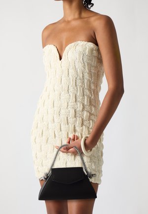 Robe crème bustier avec un design texturé et tissé ; associée à un petit sac à main noir avec une poignée chaîne et une forme angulaire.