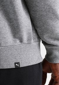 Šedá mikina s žebrovanými manžetami a lemem. Obsahuje malou černou štítek s logem Puma u lemu. Měkká, texturovaná látka.