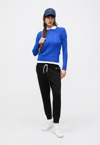 Zils atslegpītā džemperis, zem tā balta apkaklīte, melnas sašaurinātas sporta bikses, balti sporta apavi un zils cepure ar logotipu. Ikdienišķs tērps.