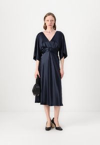 Tamsiai mėlyna satino suknelė su giliu V formos iškirpimu, apvyniojimo dizainu ir kimono rankovėmis, siekiančiomis iki vidurio blauzdų. Modelis laiko juodą rankinę ir dėvi aukštakulnius batus.