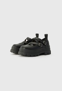 Black platform shoes with crossed straps and silver buckles. Made from smooth material with a thick, textured sole featuring a rugged tread.