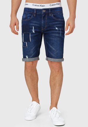 Dunkelblauje Denim-Shorts mit schäbigen Stellen und fransigem Saum. Aufgerollte Bündchen und ein Calvin Klein-Logo am Bund. Aus Baumwollmaterial.