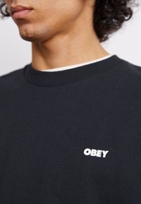 Μαύρη βαμβακερή μπλούζα με κολάρο τύπου crew και λευκό λογότυπο "OBEY" στο στήθος. Λείο ύφασμα με κοντά μανίκια.