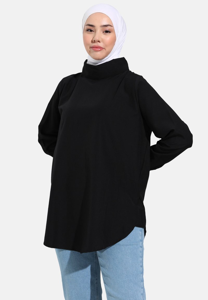 Tunique noire à manches longues avec un col haut, fabriquée en tissu doux, présente des fentes latérales et une coupe décontractée. Associée à un jean bleu clair.