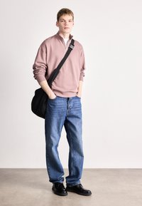 Světle růžová zipová mikina, modré džíny, černé boty a černá kabelka přes rameno. Materiál vypadá jemně a má volný střih.