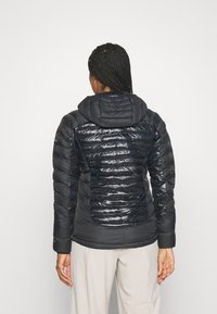 Giacca nera isolata con cappuccio, caratterizzata da una combinazione di texture lucide e opache, design trapuntato e silhouette aderente.