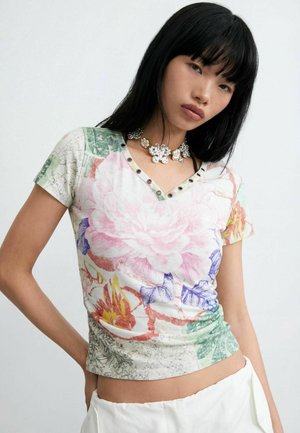 Kvinde iført en V-hals T-shirt med blomsterprint og hulmønstrede detaljer samt en sølv sommerfugl-chokerhalskæde, poserer mod en ensfarvet baggrund.