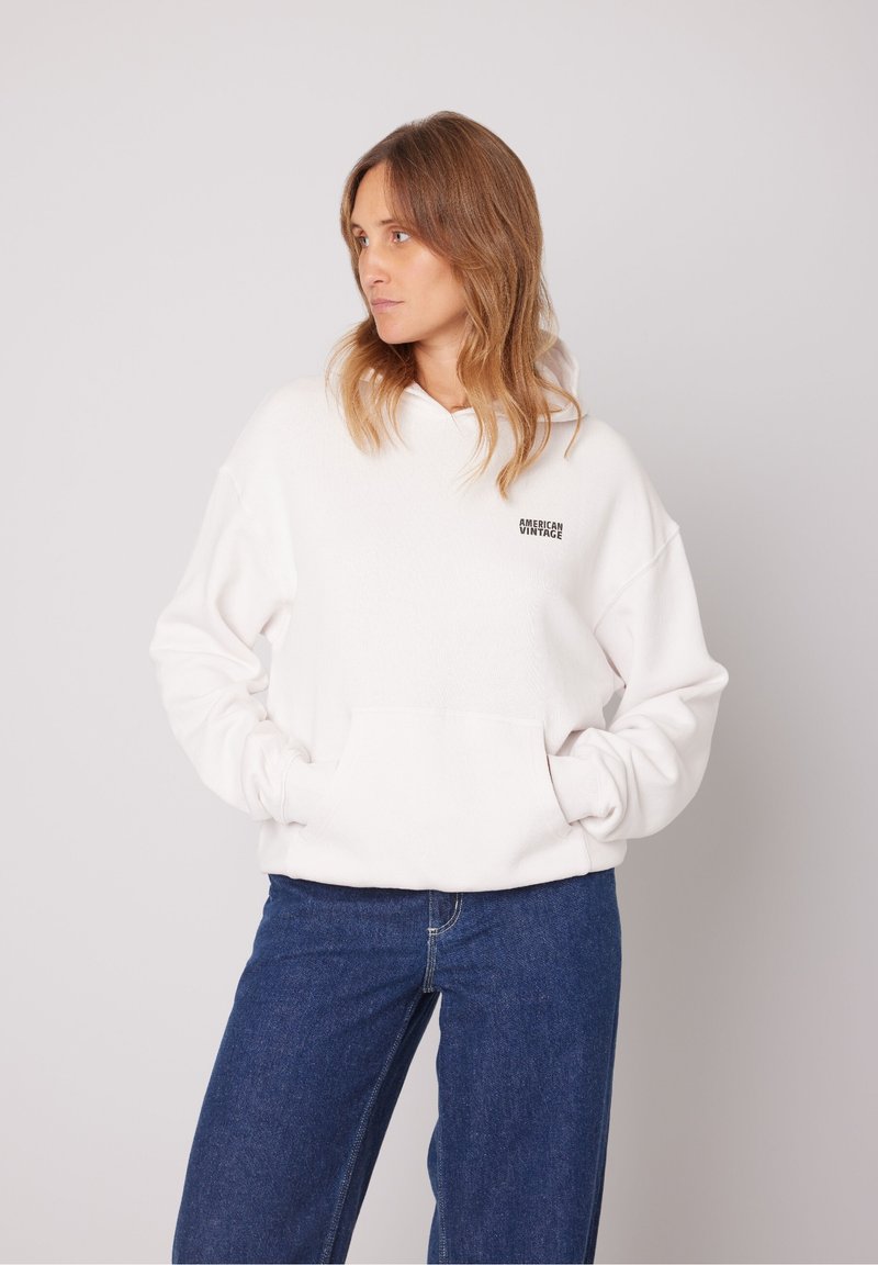 Weißer Kapuzenpullover mit weicher Textur und Fronttasche, ausgestattet mit einem kleinen schwarzen "American Vintage"-Logo auf der linken Brust.