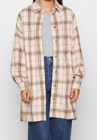 Donna che indossa una camicia button-up a quadri beige oversize sopra un top grigio e jeans blu, di fronte a uno sfondo uniforme.