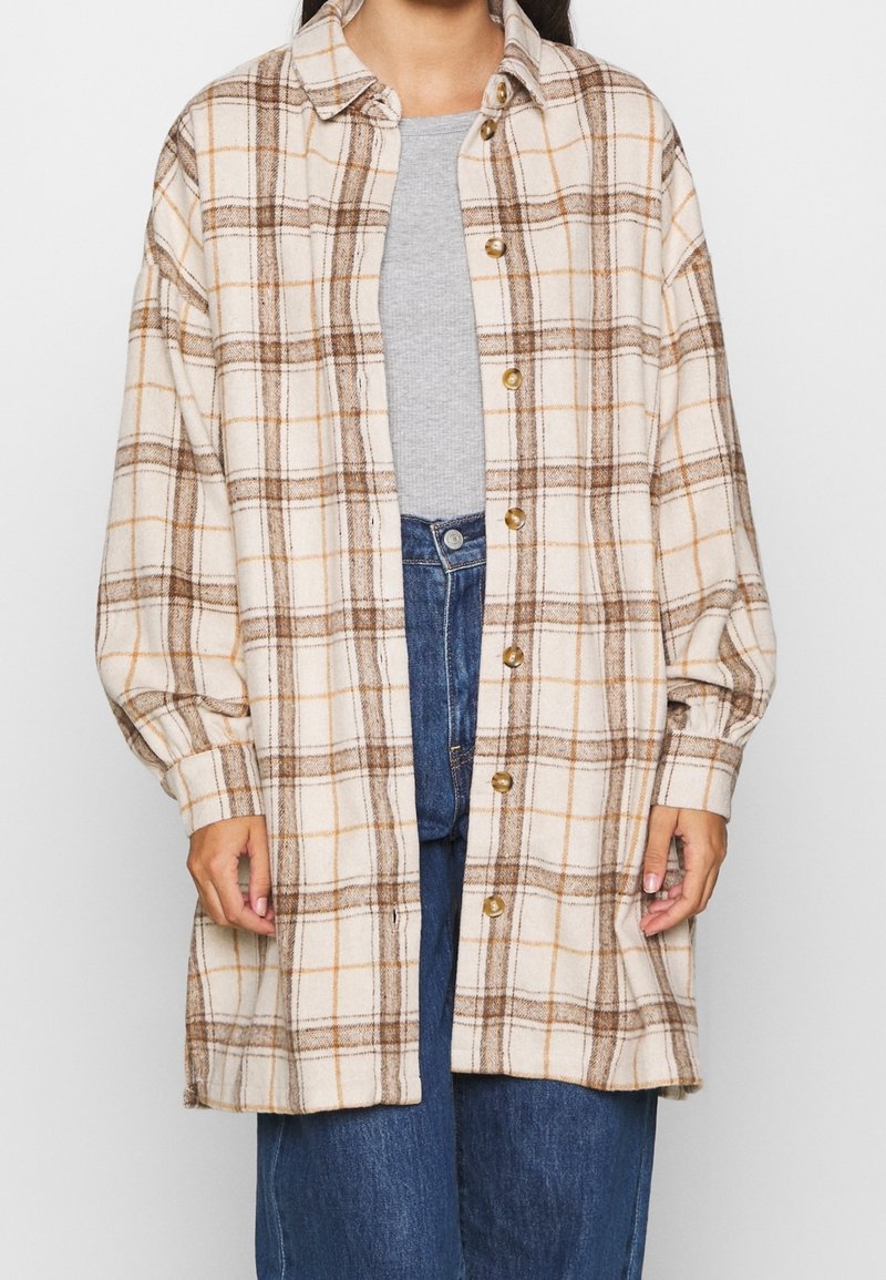 Donna che indossa una camicia button-up a quadri beige oversize sopra un top grigio e jeans blu, di fronte a uno sfondo uniforme.