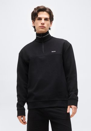 Młody mężczyzna o średniej długości włosach, ubrany w czarny pulower z krótkim suwakiem i logo "BOSS" na piersi, stoi na tle jednolitego jasnego tła.