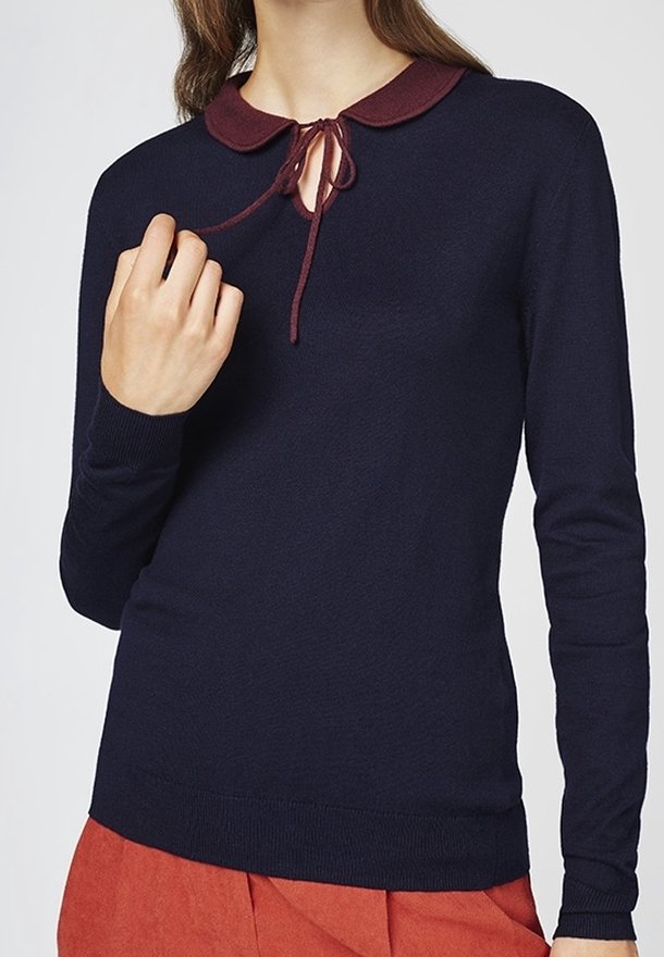 Pull bleu marine à manches longues avec un col Peter Pan bordeaux et un nœud, porté avec un pantalon couleur rouille, visible du cou aux hanches.