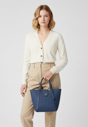 Navyblaue Ledertasche mit strukturierter Form, kurzen Griffen und Knopfverschluss, gehalten von einer Person in einer hellen Strickjacke und beigen Hosen.