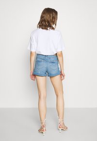 Žena stojící s výhledem od sebe, oblečená v bílém tričku s krátkým rukávem, světle modrých riflových šortkách s ohrnutými nohavicemi a bílých sandálech na podpatku.