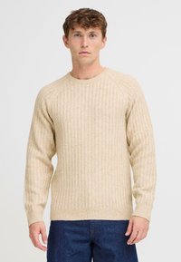 Suéter beige de canalé en un tejido texturizado, con un escote redondo y mangas largas. Combinado con jeans de color azul oscuro.