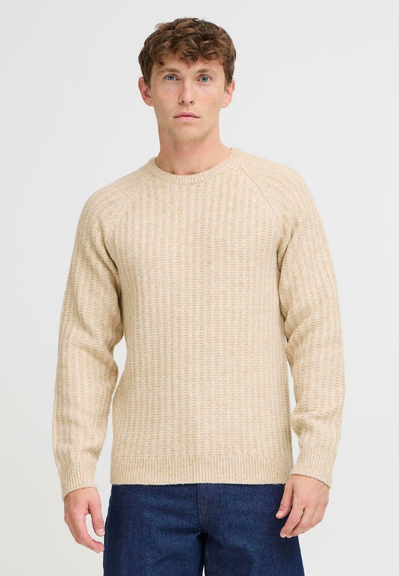 Suéter beige de canalé en un tejido texturizado, con un escote redondo y mangas largas. Combinado con jeans de color azul oscuro.