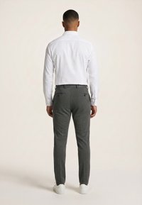 Homme debout de dos, portant une chemise blanche à manches longues, un pantalon gris foncé et des baskets blanches sur un fond uni clair.