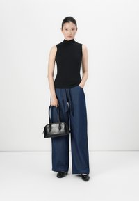 Mulher vestindo um turtleneck preto sem mangas, calças jeans azuis de perna larga, sapatilhas pretas, segurando uma bolsa preta contra um fundo branco liso.