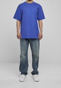 Niebieska bawełniana koszulka z krótkim rękawem, w połączeniu z jasnoniebieskimi, wypłowiałymi dżinsami. Białe trampki dopełniają outfit, stojąc na szarym tle.