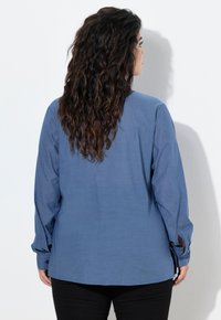 Camicia blu a maniche lunghe con una vestibilità comoda, polsini con bottoni e una texture liscia, abbinata a pantaloni neri. La vista posteriore non presenta elementi di design.