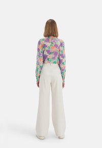 Blouse à fleurs dans des tons de violet, vert et orange, à manches longues et avec des poignets élastiques, associée à un pantalon crème à jambes larges. Vue arrière affichée.