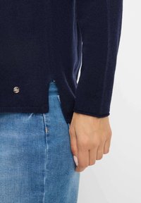 Hand in hand, gekleed in een donkerblauw vest met zijsplit en metalen embleem, in combinatie met blauwe denim jeans.