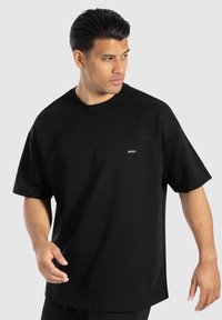 Schwarzes, übergroßes T-Shirt aus Baumwolle mit Rundhalsausschnitt, kurzen Ärmeln und kleinem Logo auf der Brust. Glatte Textur.