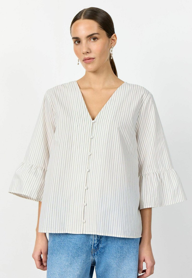 Vrouw met donker haar naar achteren gebonden, draagt hangende oorbellen, een witte blouse met dunne verticale strepen, knoopjes aan de voorkant en uitlopende driekwart mouwen, gecombineerd met een blauwe spijkerbroek.