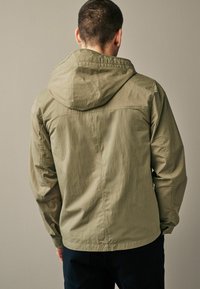 Jachetă ușoară, de culoare verde măslină, cu glugă, textură netedă, manșete elastice și un design simplu. Vederea din spate arată cusături plate și detalii minime.