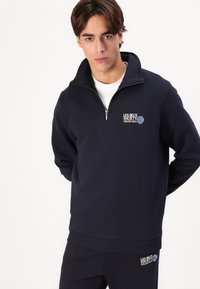 Felpa marina con colletto zip, orlo e polsini a coste. Presenta del testo del logo bianco sul petto e dettagli grafici blu, realizzata in tessuto morbido.