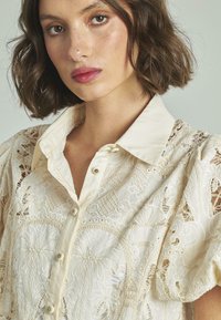 Weiße Spitzenbluse mit kragenförmigem Ausschnitt, durchgehender Knopfleiste und aufwendigen bestickten Mustern. Sie hat Puffärmel und strukturierte Details.