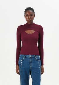 Bordeaux ribbed longsleeve top met een hoge hals en een uitsnede bij de borst, gecombineerd met blauwe jeans met studdecoraties.