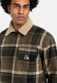 Mand iført en grøn og brun ternet jakke med beige sherpa-krave og brystlomme med Quiksilver-logo-patch.