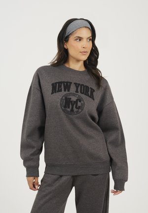 Donna che indossa un completo felpa e pantaloni grigio scuro con la scritta "NEW YORK NYC" stampata sul davanti, in posa contro uno sfondo bianco semplice.