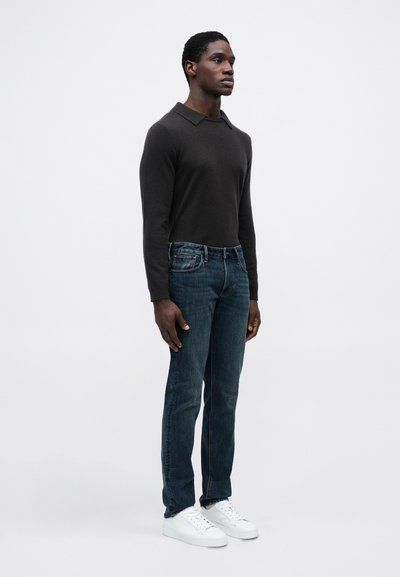 Fiatal férfi állva, sötét hosszúujjú pólót, kék farmert és fehér sportcipőt visel egy sima világos háttér előtt.