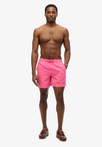 Costume da bagno rosa con vita elasticizzata e cordino. Presenta un piccolo logo sulla gamba sinistra. Indossato con scarpe da barca marroni.
