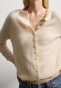 Cardigan in misto lana crema con collo rotondo, otto bottoni rivestiti in tessuto, polsini e orlo a coste, caratterizzato da una morbida e pelosa texture.