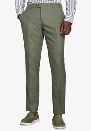 Uomo che indossa pantaloni su misura color verde oliva, camicia a righe e mocassini in camoscio verde oliva abbinati con suola bianca, in piedi con una mano in tasca.